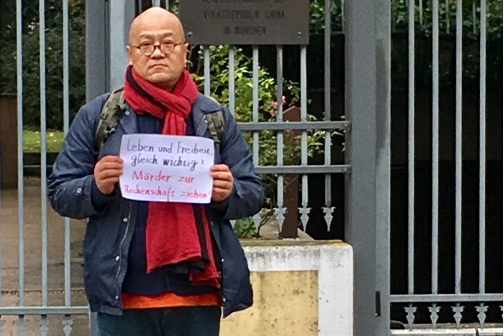 楊偉東站在慕尼黑中國大使館辦事處前面，拿著標語，上面寫著「生命與自由一樣重要，謀殺者必得清算」。（作者提供）
